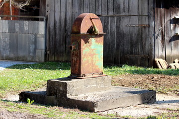 Obraz premium Large vintage retro partially rusted public hand water pump mounted on dilapidated concrete foundation surrounded with grass and wooden barn in background on warm sunny spring day