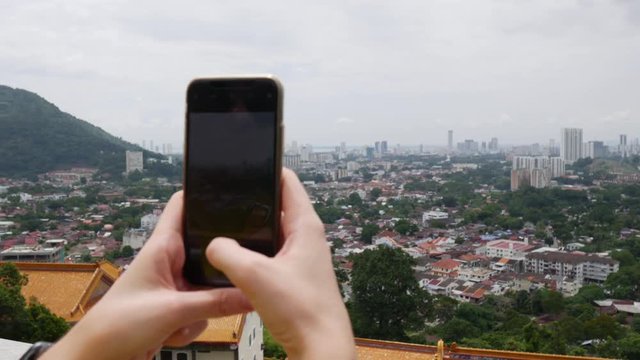 Young woman makes vertical photos of city on the smartphone standing on the mountain. Camera over the shoulder. Cloudy sky. Closeup. 4k
