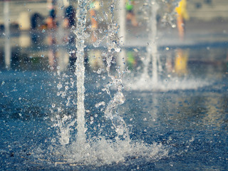 Close up view on water streams beating out of stainless grate of dry fountain. Water drops splashing on granite coating. Town square in summer. Selective soft focus. Blurred background