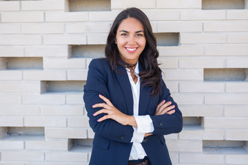 Confident cheerful businesswoman. Content young businesswoman standing with crossed arms near brick...