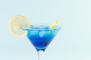 Cool summer blue beverage margarita for party with ice cubes,  lemon slice, yellow straw on white wood board and mint color wall, top part, details.