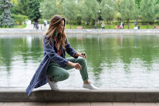 young Caucasian girl, natural appearance, long hair, beautiful face, green pants, pink shirt, blue long jacket, white sneakers. Sitting by side lake in city park. linen womens clothing.