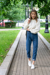 Beautiful European teen girl walking on pavement in city green park. Happy smile caucasian girl. Lifestyle, pullover, jeans, sneakers. It is interesting pose full length in fashionable look.