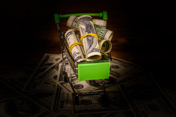 Small shopping cart with rolled up one hundred dollar banknotes on a wooden background