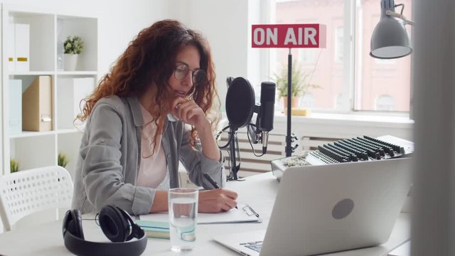 Pan shot of young Caucasian woman sitting at desk in broadcast studio, making notes in document and then typing on laptop keyboard, taking off eyeglasses, touching her hair and continuing writing