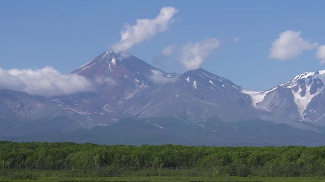 Zoom in view of cone of active Avachinsky Volcano, alpine lake, clouds drifting across blue sky near mountains. Summer landscape of Kamchatka Peninsula, popular travel destinations in Russian Far East