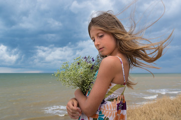 The wind develops the hair of a beautiful girl with a bouquet of wild flowers against the background of the sea and clouds