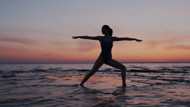 Young sport and flex woman exercising yoga, doing warrior pose on beach during sundown sky. Silhouette of a Slim and fit girl performing kundalini asana outdoor.