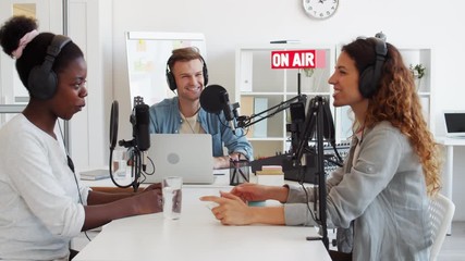 Medium shot of three diverse radio hosts sitting at microphones at table in studio and having pleasant talk on air