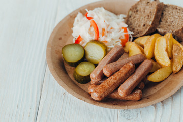 Tasty sausages with fried potatoes, cucumber, cabbage and chlobe in a paper plate on a wooden table.