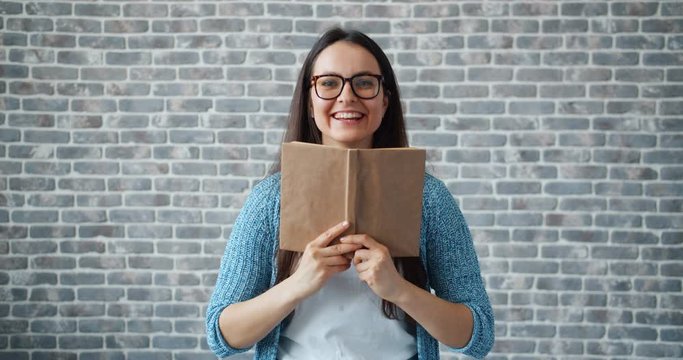 Portrait of happy girl in glasses reading book hiding face then smiling standing on brick wall background. Youth culture, education and lifestyle concept.