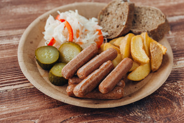 Tasty sausages with fried potatoes, cucumber, cabbage and chlobe in a paper plate on a wooden table.