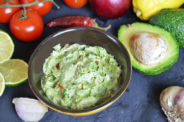 Bowl of freshly prepared guacamole. Ingredients on the background tomatoes, hot pepper, lime, avocado. Guacamole nachos and guacamole ingredients on dark wooden background.