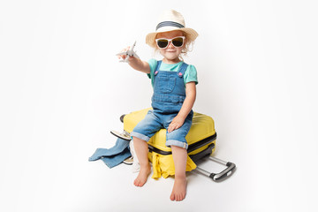 Funny curly little baby girl in a hat, T-shirt and jeans with a yellow suitcase is smiling on white background waiting for an airplane. concept trip with kids.