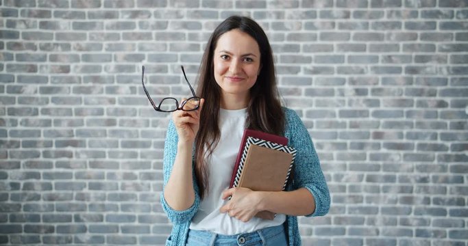Young beautiful woman is taking off glasses holding book standing on brick wall background smiling. People, education and youth lifestyle concept.