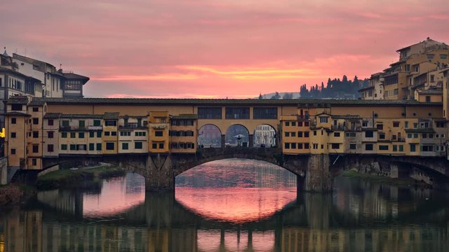 Florence, Italy. Yellow Ponte Vecchio Bridge against pink and orange sunset sky. Sky is reflecting in Arno River. Panning shot, UHD