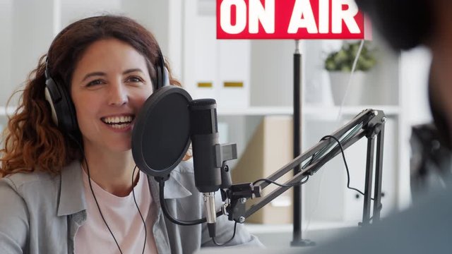 Back view of female Caucasian radio host with long curly hair sitting at mike, communicating cheerfully with man sitting in front of her