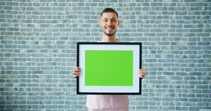 Portrait of bearded man holding green chroma key mockup picture and smiling standing on brick wall background. People, greenscreen and emotions concept.