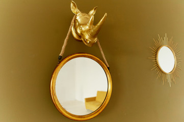 A round mirror on a brown and gold wall. Hanging on the fore head of a rhinoceros, painted in gold color. Interior design