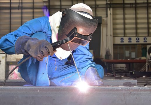 Welder is assembling the workpiece by process shielded metal arc welding (SMAW). Welder in blue uniform, safety shoes, leather gloves, welding mask. He is sitting and welding.