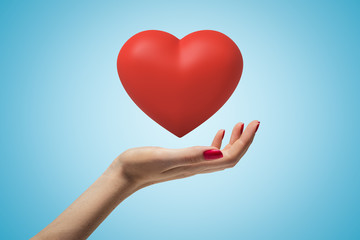 Side closeup of woman's hand facing up and levitating beautiful red heart on light-blue gradient background.