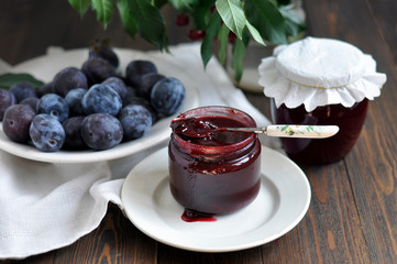 Glass jar of jam and ripe plums on a dark wooden table.
