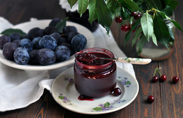Glass jar of jam and ripe plums on a dark wooden table.