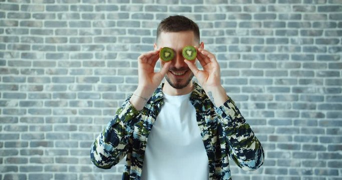 Portrait of handsome young man with kiwi fruit on eyes smiling standing on brick wall background having fun. Healthy food, modern lifestyle anf youth concept.