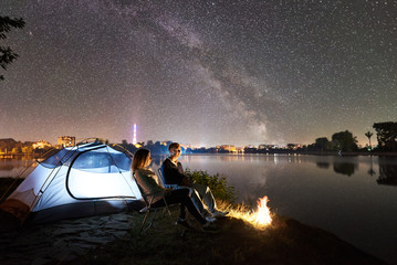 Night camping on lake shore. Couple tourists - man and woman sitting on chairs near campfire and...