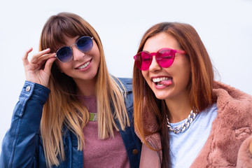 Photo of two charming brunette female friends hugging each other, showing peace sign, looking at camera on city street