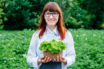 Beautiful female farmer holding green cabbage leaves in wicker basket, smiling at the camera and surrounded by plenty of plants in her vegetable garden