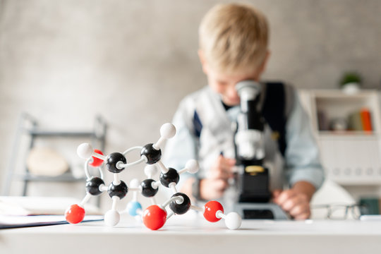 Plastic Model Of Atoms And Molecules In The Foreground. A Young Boy In A White Shirt And Sweater Examines The Sample Under A Microscope