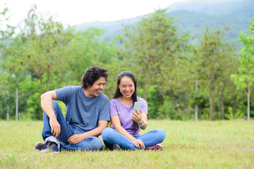 Happy Asian couple sitting on green grass in outdoor and laughing with smartphone