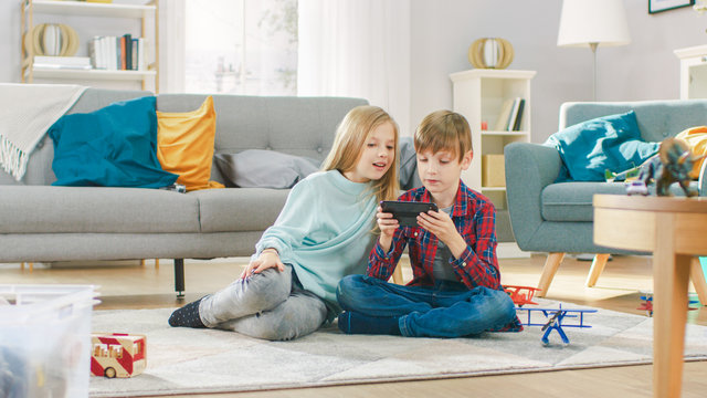 At Home: Cute Little Girl and Sweet Boy Playing Video Game on a Smartphone, Boy Holds Mobile Phone in Horizontal Landscape Mode. Children Playing in Online Videogame Sitting on a Carpet.