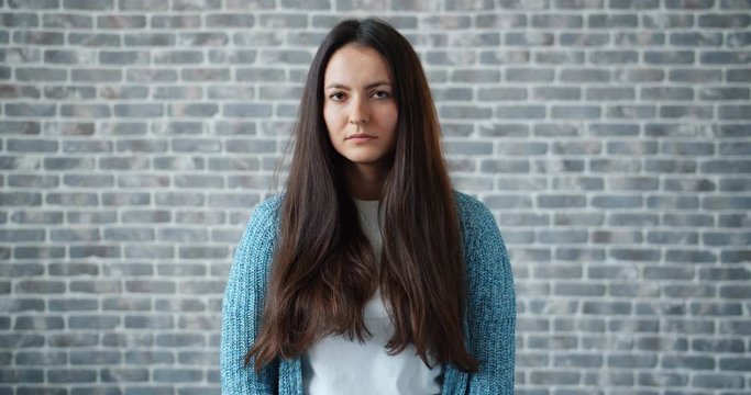 Portrait of sad young woman looking around then at camera standing alone on gray brick wall background. Human emotions, upset youth and people concept.