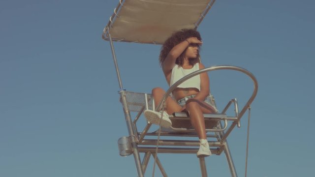 mixed race black young woman outdoors, summer sunset light, mixed race woman sits on the lifeguard chair and watching around