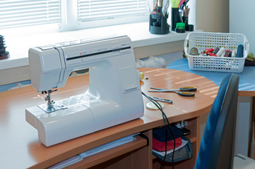 white sewing machine, basket with threads on wooden table and blue chair near window in sewing studio