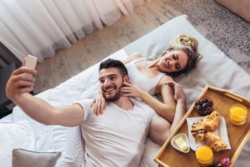 Young happy couple having breakfast in luxury hotel room using smart phone to take selfie photo.