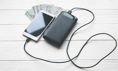 Black power bank with usb and smartphone on wooden background. Dollar notes lying nearby. Modern information technology photo.
