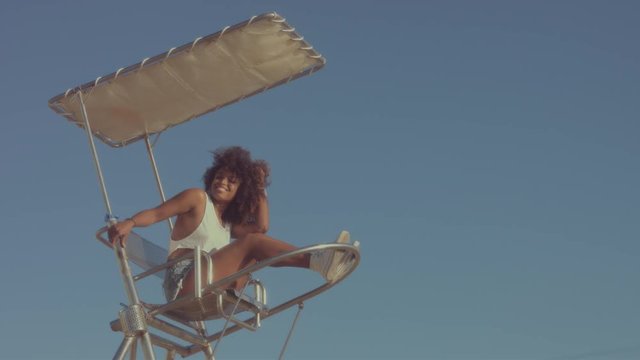 mixed race black young woman outdoors, summer sunset light, mixed race woman sits on the lifeguard chair and makes faces have fun slow motion from 60 fps