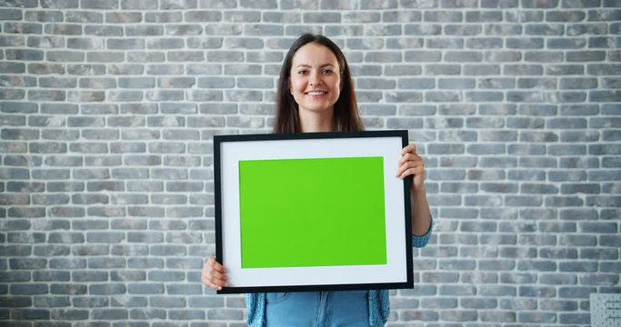 Portrait of beautiful girl holding green chroma key mockup picture and smiling standing on brick wall background. People, green screen and emotions concept.