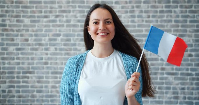 Portrait of smiling pretty lady holding French flag on brick wall background standing alone with happy face and hair flying in wind. Youth and travelling concept.