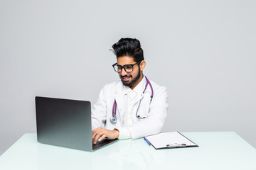 Male indian doctor wearing a white coat, sitting at a desk working with his laptop.