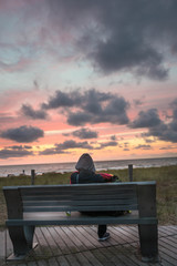 Naklejka na ściany i meble Jemand sitzt und schaut den Sonnenuntergang auf einer Sitzbank am Meer am Strand