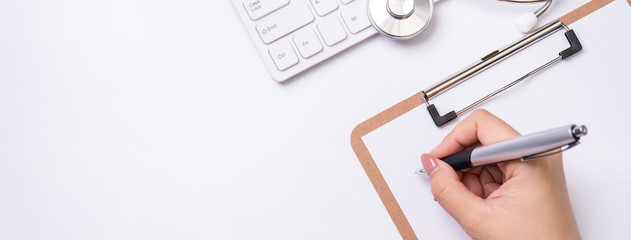 Female doctor writing a medical record case over clipboard on white working table with stethoscope,...
