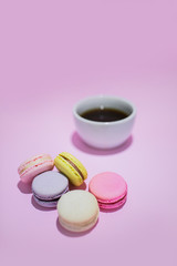 Overview of sweet colorful macaroon biscuits lying on pastel pink background. A mug with black coffee beside.
