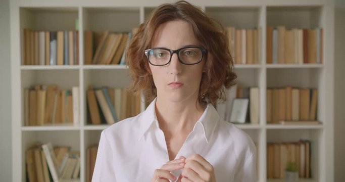 Closeup portrait of adult redhead caucasian businesswoman in glasses looking at camera indoors with bookshelves on the background