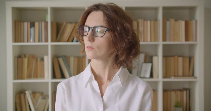 Closeup portrait of adult redhead attractive caucasian businesswoman in glasses looking at camera indoors with bookshelves on the background