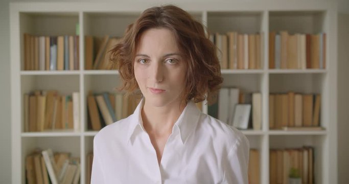 Closeup portrait of adult redhead attractive caucasian businesswoman looking at camera indoors with bookshelves on the background