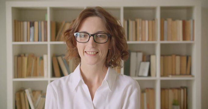 Closeup portrait of adult redhead attractive caucasian businesswoman looking at camera smiling happily indoors with bookshelves on the background